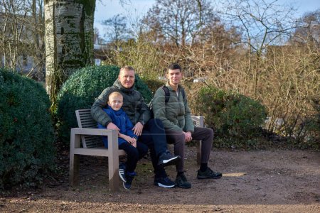 Dad with two sons sitting on a bench in autumn park. Experience the tranquility of familial bonds in the heart of autumn with this serene image. A father, 40 years old, and his two sons - a beautiful