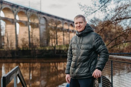 Élégance intemporelle : Homme de 40 ans veste élégante au bord de la rivière Neckar et pont historique à Bietigheim-Bissingen, Allemagne. Découvrez l'attrait des saisons comme un homme charismatique de 40 ans se tient