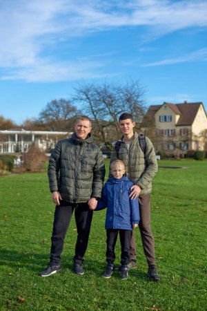Armonía familiar: padre, 40 años y dos hijos: hermoso niño de 8 años y joven de 17 años, parado en el césped en un parque con edificios de madera mestiza vintage, Bietigheim-Bissingen
