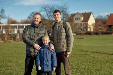 Familienharmonie: Vater, 40 Jahre alt, und zwei Söhne - ein schöner 8-jähriger Junge und ein 17-jähriger junger Mann stehen auf dem Rasen in einem Park mit historischen Fachwerkhäusern, Bietigheim-Bissingen