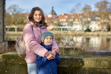 Riverside Family Harmony: Mother, 30 Years Old, and Son - Beautiful 8-Year-Old Boy, Standing by Neckar River and Historic Half-Timbered Town, Bietigheim-Bissingen, Germany, Autumn. Sumérgete en