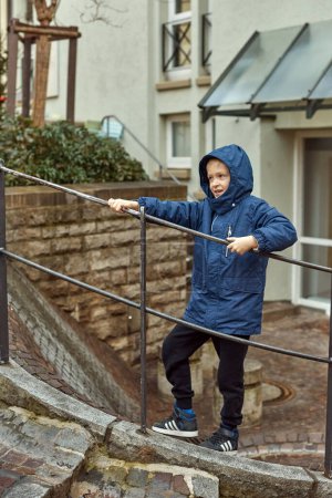 Joyful Boy in Blue Winter Jacket on Outdoor Stairs with City Buildings in the Background, Autumn or Winter. Experience the delight of the season with this heartwarming image featuring a cheerful 8