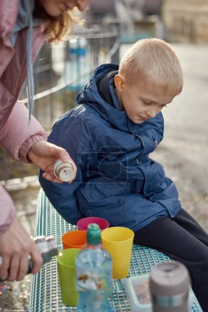 Familien-Picknick-Vergnügen: Fröhlicher 8-jähriger blonder Junge in blauer Winterjacke sitzt auf Bank, während Mama Tee aus Thermoskannen gießt, Herbst oder Winter. Tauchen Sie ein in die Wärme familiärer Momente mit diesem