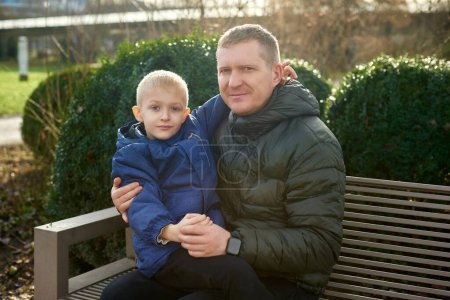 Autumnal Family Affection: Father, 40 Years Old, and Son - Beautiful 8-Year-Old Boy, Seated in the Park. Embrace the warmth of familial love in the autumnal air with this heartwarming image. A father