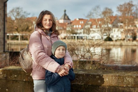 Riverside Family Harmony: Mother, 30 Years Old, and Son - Beautiful 8-Year-Old Boy, Standing by Neckar River and Historic Half-Timbered Town, Bietigheim-Bissingen, Germany, Autumn. Plongez dans