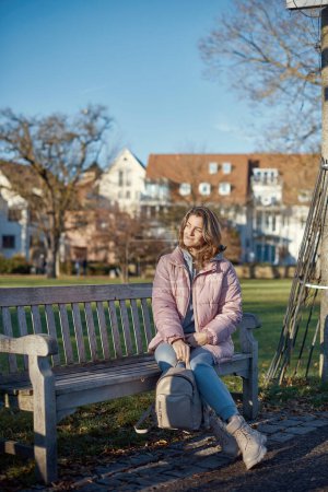 Embrace the tranquility of winter with this captivating image featuring a beautiful girl in a pink winter jacket, leisurely sitting on a bench in a park against the backdrop of a charming old European