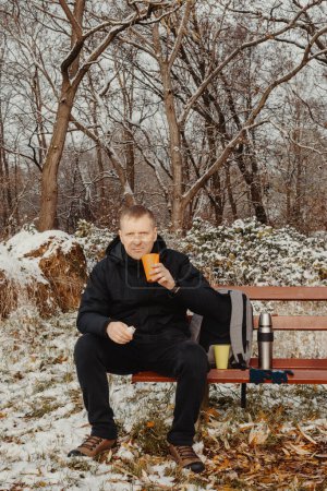 Winterliche Gelassenheit: 40-jähriger Mann genießt Tee auf schneebedeckter Bank im Landschaftspark. Tauchen Sie ein in die ruhige Schönheit des Winters, als ein 40-jähriger Mann Trost auf einer verschneiten Bank in einem ländlichen Park findet