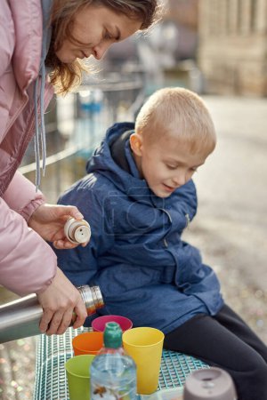 Familien-Picknick-Vergnügen: Fröhlicher 8-jähriger blonder Junge in blauer Winterjacke sitzt auf Bank, während Mama Tee aus Thermoskannen gießt, Herbst oder Winter. Tauchen Sie ein in die Wärme familiärer Momente mit diesem