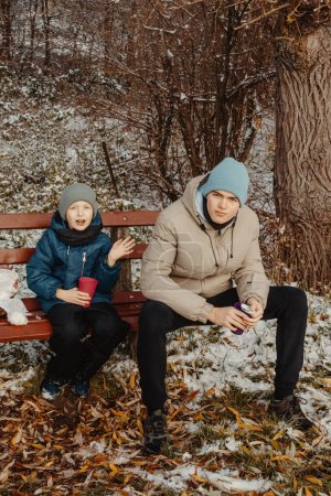 Capture the heartwarming essence of winter as two brothers, aged 8 and 17, share a special moment on a snow-covered bench in a serene rural park. Sipping hot tea from a thermos, they immerse