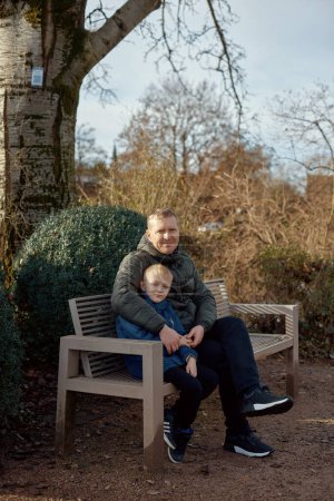 Herbstliche Zuneigung in der Familie: Vater, 40 Jahre alt, und Sohn - ein wunderschöner 8-jähriger Junge, der im Park sitzt. Umarmen Sie die Wärme der familiären Liebe in der herbstlichen Luft mit diesem herzerwärmenden Bild. Ein Vater
