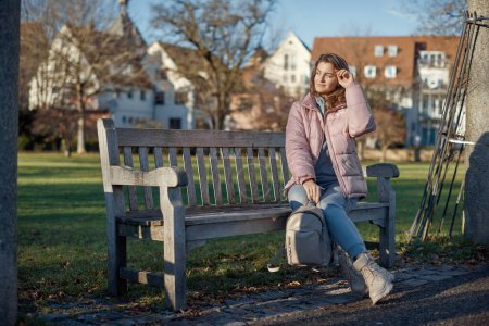 Experience the festive spirit of winter with this delightful image capturing a beautiful girl in a pink winter jacket sitting on a bench in a park, set against the backdrop of the historic town of