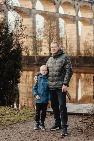 Serenidad familiar: Hombre guapo de 40 años e hijo de 8 años en medio de la belleza del río Neckar y el puente histórico, Bietigheim-Bissingen, Alemania, invierno u otoño. Abrace el calor de los lazos familiares