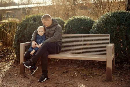 Herbstliche Zuneigung in der Familie: Vater, 40 Jahre alt, und Sohn - ein wunderschöner 8-jähriger Junge, der im Park sitzt. Umarmen Sie die Wärme der familiären Liebe in der herbstlichen Luft mit diesem herzerwärmenden Bild. Ein Vater