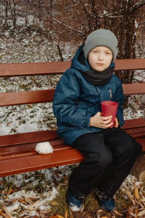 Winter-Picknickvergnügen: 8-jähriger Junge genießt Tee auf verschneiter Bank im Landschaftspark Erleben Sie den Zauber des Winters durch die Linse dieses fesselnden Fotos, auf dem ein 8-jähriger Junge eine