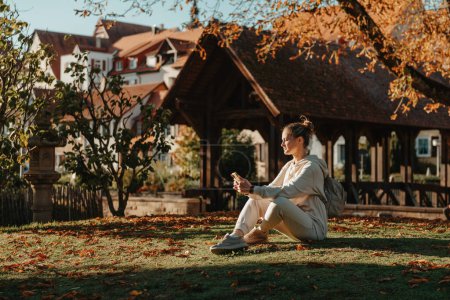 Photo for Young fashionable teenage girl with smartphone in park in autumn sitting at smiling. Trendy young woman in fall in park texting. Retouched, vibrant colors. Beautiful blonde teenage girl wearing casual - Royalty Free Image