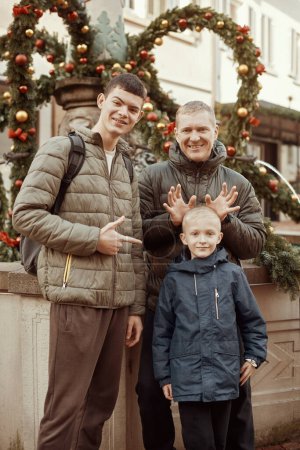 Joyful Family Portrait: Father and Two Sons by Festive Vintage Fountain (en inglés). Captura la esencia de la felicidad familiar con esta imagen conmovedora con un padre guapo con sus dos hijos de pie