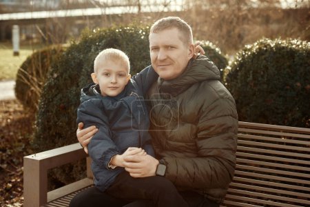 Autumnal Family Affection: Father, 40 Years Old, and Son - Beautiful 8-Year-Old Boy, Seated in the Park. Embrace the warmth of familial love in the autumnal air with this heartwarming image. A father