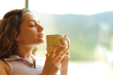 Téléchargez les photos : Femme heureuse sentant le café relaxant à côté d'une fenêtre à la maison avec les yeux fermés - en image libre de droit