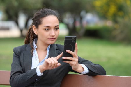 Téléchargez les photos : Femme d'affaires assise sur un banc dans un parc en utilisant le téléphone - en image libre de droit