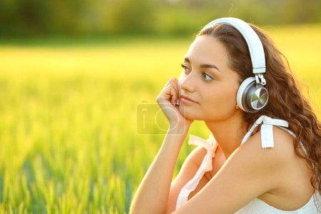 Foto de Mujer pensando escuchando música mirando lejos en un campo al atardecer - Imagen libre de derechos