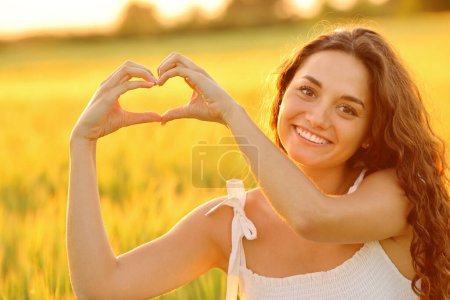 Téléchargez les photos : Femme heureuse faisant forme de coeur avec les mains au coucher du soleil dans un champ de blé - en image libre de droit