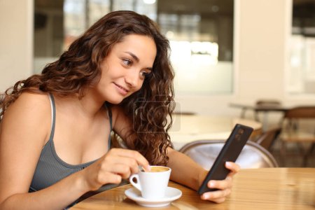 Foto de Mujer revisando el teléfono inteligente y revolviendo café sentado en un bar - Imagen libre de derechos