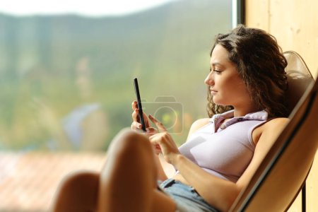 Photo for Woman using phone sitting on a chair at home beside a window - Royalty Free Image