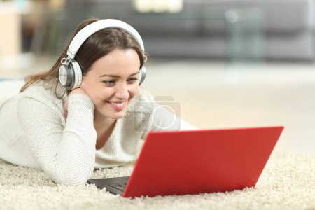 Téléchargez les photos : Joyeux adolescent portant des écouteurs regardant les médias sur ordinateur portable sur le sol à la maison - en image libre de droit