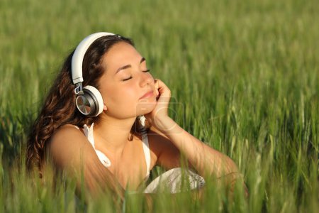 Foto de Mujer relajada escuchando música usando auriculares en un campo de cebada verde - Imagen libre de derechos