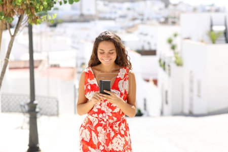 Foto de Front view portrait of a happy tourist using smart phone walking in a white spanish town - Imagen libre de derechos