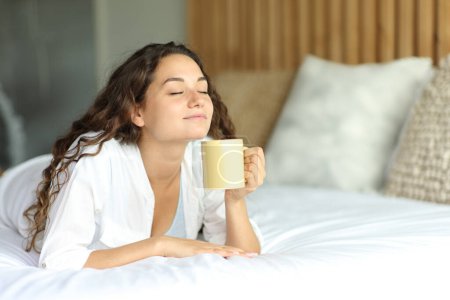 Foto de Happy woman lying on a bed enjoying a cup of coffee - Imagen libre de derechos