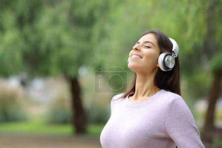 Téléchargez les photos : Happy woman breathing fresh air with headphones on green background in a park - en image libre de droit