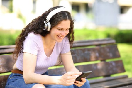 Téléchargez les photos : Woman with headphones watching content on smartphone siting on a bench - en image libre de droit