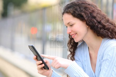 Téléchargez les photos : Happy woman vérifie le contenu du téléphone intelligent assis dans la rue - en image libre de droit