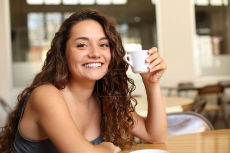 Foto de Mujer feliz sosteniendo taza de café mirándote sentado en un bar y sonriendo - Imagen libre de derechos