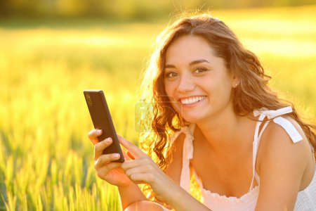 Téléchargez les photos : Femme heureuse posant dans un champ tenant un téléphone portable au coucher du soleil - en image libre de droit