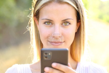 Photo for Front view portrait of a beautiful woman holding phone looking at camera outdoors - Royalty Free Image