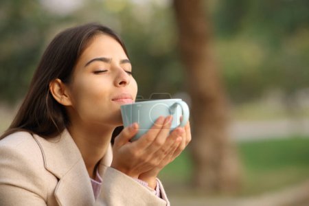 Foto de Mujer en invierno oliendo aroma a café sentada en un parque - Imagen libre de derechos