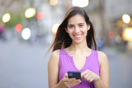 Téléchargez les photos : Portrait d'une femme heureuse tenant un téléphone intelligent dans la rue vous regardant - en image libre de droit