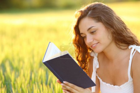 Téléchargez les photos : Femme heureuse assise dans un champ lisant un livre de papier au coucher du soleil - en image libre de droit