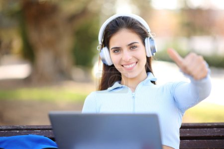 Téléchargez les photos : Étudiant heureux avec ordinateur portable et casque avec pouces sur un banc dans un parc - en image libre de droit