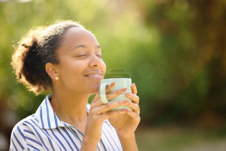 Téléchargez les photos : Femme noire détendue sentant tasse de café reposant dans un parc - en image libre de droit