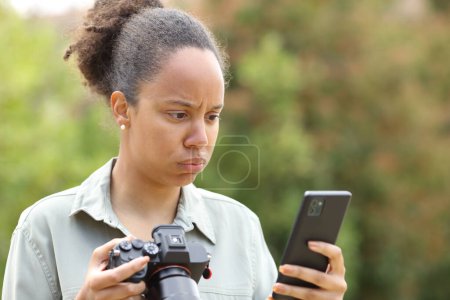 Téléchargez les photos : Photographe noir frustré tenant un appareil photo sans miroir vérifiant le téléphone dans un parc - en image libre de droit