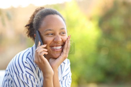 Téléchargez les photos : Heureuse femme noire franche appelant au téléphone dans un parc - en image libre de droit