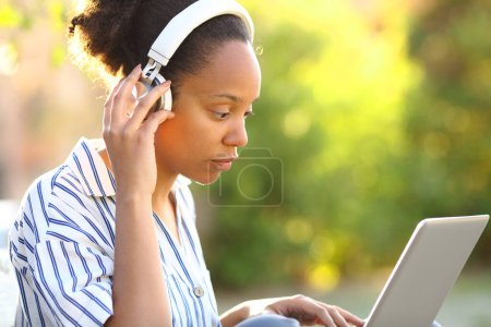 Photo for Serious black woman wearing headphone watching video on laptop in a park - Royalty Free Image
