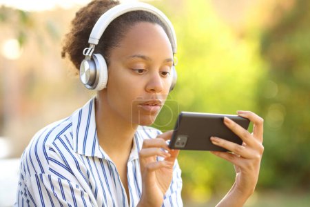 Téléchargez les photos : Femme noire sérieuse portant un casque regarder des vidéos sur le téléphone dans un parc - en image libre de droit