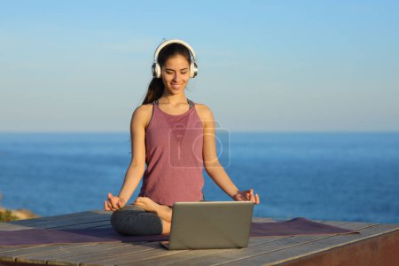 Photo for Yogi doing yoga watching video tutorial online on laptop on the beach - Royalty Free Image