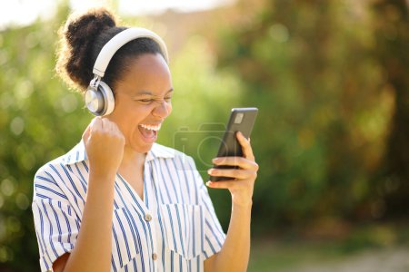 Foto de Mujer negra emocionada usando auriculares celebrando escuchar música con teléfono en un parque - Imagen libre de derechos