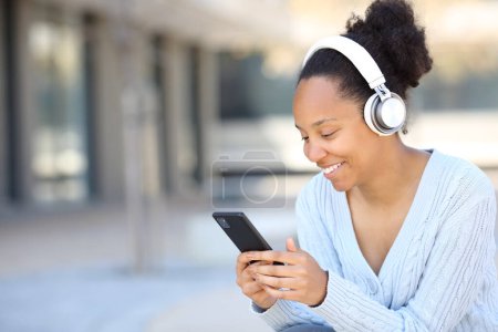 Téléchargez les photos : Heureuse femme noire écoutant de la musique avec casque dans la rue à l'aide d'un téléphone intelligent - en image libre de droit