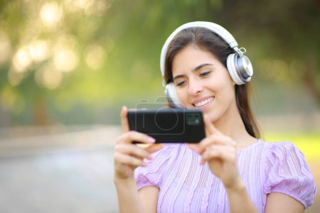 Téléchargez les photos : Femme heureuse avec casque regarder des vidéos sur le téléphone dans un parc - en image libre de droit